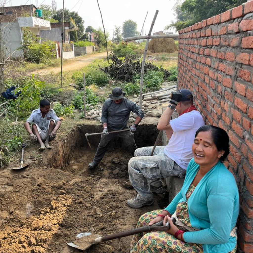volunteer digging hole while local woman looks on laughing Nepal